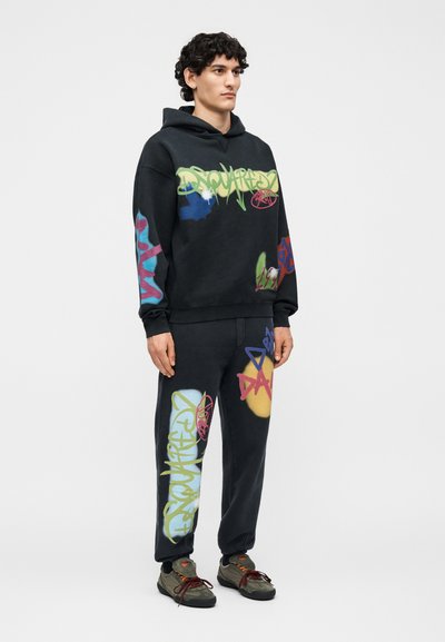 Νέος άνδρας στέκεται, φοράει μαύρη φούτερ με κουκούλα και φόρμες με πολύχρωμα σχέδια στυλ γκράφιτι και ελιά πράσινα αθλητικά με κόκκινα κορδόνια.