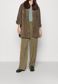 Ruskea corduroy-ylisuuri paitajakku, kevyt vihreä toppi, oliivinvihreät leveälahkeiset housut ja tummanvihreät kengät liukkaalla pinnalla.