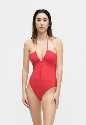 Γυναίκα φοράει κόκκινο ολόσωμο μαγιό με λαιμόκοψη halter και κορδόνι μπροστά, στέκεται μπροστά σε ένα απλό φωτεινό φόντο.