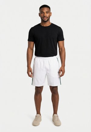 Uomo in piedi di fronte, che indossa una maglietta nera a maniche corte, pantaloncini sportivi bianchi con strisce verdi e scarpe da ginnastica beige su sfondo bianco.