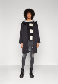 Black waterproof jacket with a hood, layered over a black and cream striped sweater. Gray leggings and black lace-up boots complete the look.