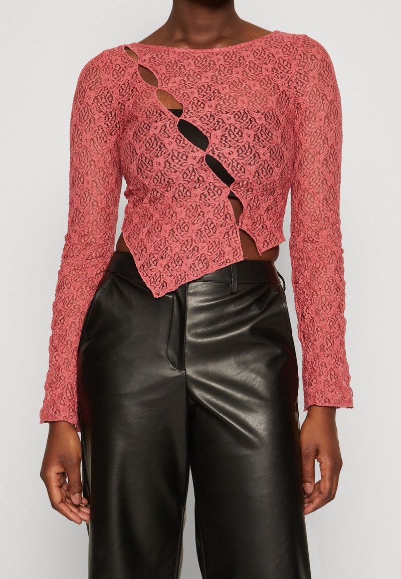 Pink lace long-sleeve top featuring a cut-out design with a curved hem, paired with high-waisted black leather pants. Textured and detailed.