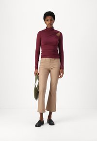 Haut bordeaux à manches longues avec un col montant et une découpe sur le côté, associé à un pantalon beige évasé et des mocassins noirs, portant un sac vert.