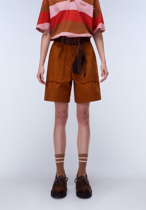 Person iført brune shorts med høj talje og store lommer, en flerfarvet stribet skjorte, brun bælte med nøglering, brune sko og sokker.