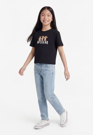 Uśmiechnięta dziewczyna z długimi czarnymi włosami, ubrana w czarną koszulkę Moschino z misiami, jasnoniebieskie dżinsy i białe trampki, stojąca na białym tle.