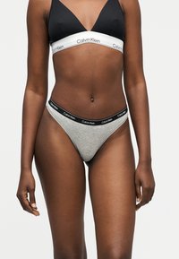 Μαύρο και άσπρο bralette της Calvin Klein με γκρι σλιπ Calvin Klein σε ένα σκουρόχρωμο άτομο που στέκεται μπροστά σε λευκό φόντο.