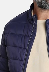 Chaqueta de plumas azul marino con cierre de cremallera, diseño acolchado y una textura suave, llevada sobre un suéter beige.