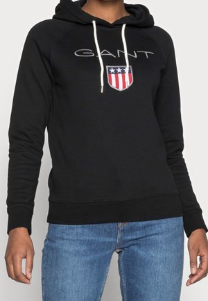 Sort hættetrøje med hvide snore, "GANT" tekst og rød, hvid og blå skjoldemblem båret med blå jeans på en person.