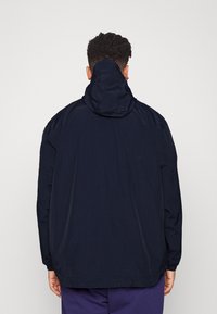 Hombre con cabello rizado corto que lleva una chaqueta con capucha suelta de color azul marino oscuro y pantalones morados, de espaldas sobre un fondo claro y liso.