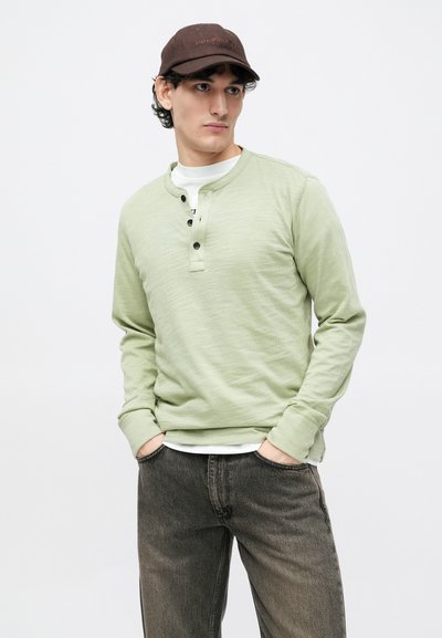 Light green long-sleeve henley shirt with three buttons, paired with dark, slightly faded jeans. Soft texture; casual fit. Brown cap.