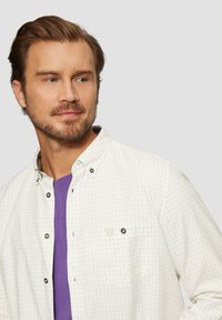 Hellbeige kariertes Hemd mit Button-Down-Kragen, ausgestattet mit einer Brusttasche und grauen Knöpfen, über einem lila T-Shirt getragen.