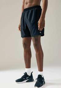 Pantaloni scurți sport negri cu un elastic la talie și buzunar lateral. Purtați cu adidași negri și șosete albe, evidențiind un design sportiv.