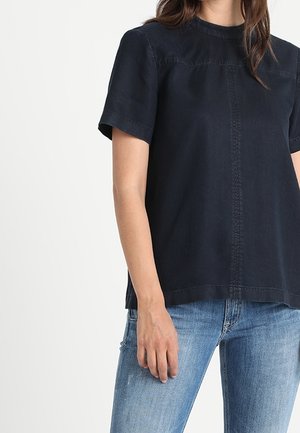 Vrouw draagt een zwart T-shirt met korte mouwen en een centrale naaddetail, gecombineerd met lichtblauwe spijkerbroek, staand tegen een effen achtergrond.