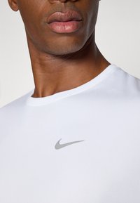 White Nike t-shirt with a round neckline, featuring a subtle silver logo on the chest. The fabric appears smooth and fitted.