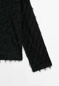 Vêtement noir à manches longues avec des motifs diagonaux texturés et à franges sur le tissu, présenté sur un fond blanc.