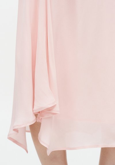 Tissu rose clair translucide s'écoulant sur les jambes, mettant en valeur un ourlet asymétrique drapé et une texture en couches.