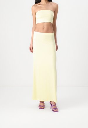 Vrouw draagt een lichtgeel strapless bandeau topje en bijpassende lange rok met paarse hoge hakken op een witte achtergrond.