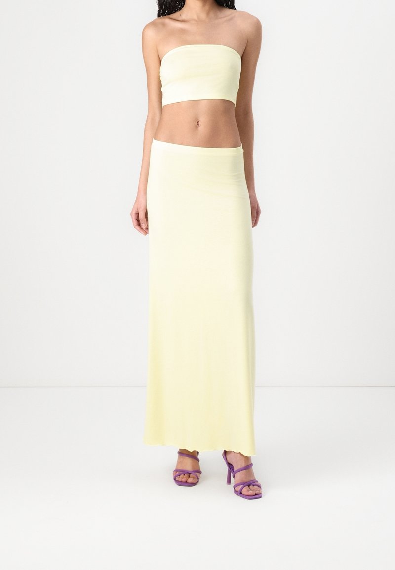 Femme portant un top bandeau jaune clair sans bretelles et une longue jupe assortie avec des sandales à talons violettes sur fond blanc.