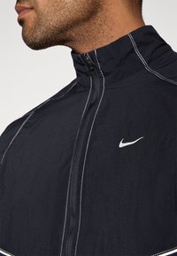 Črno jakno Nike z visokim ovratnikom, z belo šivom in majhnim logotipom na levi strani. Lahka tkanina, z zadrgo za zapiranje.