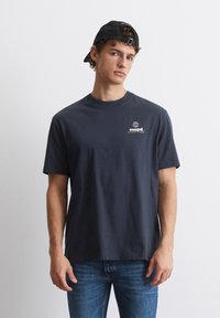 Marine bomull T-skjorte med rund hals, korte ermer, og en globuslogo trykket i hvitt og oransje på venstre bryst. Brukt sammen med blå jeans.
