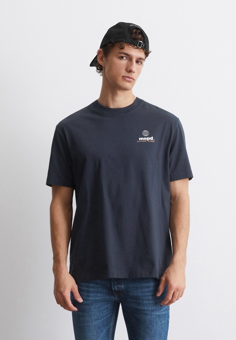 Marine bomull T-skjorte med rund hals, korte ermer, og en globuslogo trykket i hvitt og oransje på venstre bryst. Brukt sammen med blå jeans.