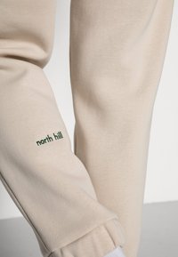 Beige cotton sweatpants with elastic cuffs, featuring green "north hill" embroidery on the left leg. Smooth texture, relaxed fit.
