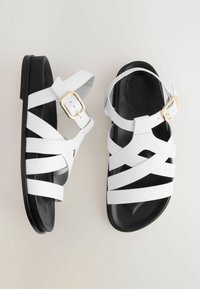 Sandali bianchi con cinturini, con una suola nera piatta, presentano un dettagli in metallo dorato e una superficie liscia. I cinturini sono larghi e intrecciati.