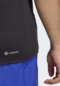Close-up of a person’s midsection wearing a black Aeroready sports shirt and blue shorts, with their right hand relaxed by their side.