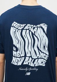 Άτομο που φοράει μπλε σκούρο πουκάμισο με παραμορφωμένο λευκό κείμενο "New Balance", τη φράση "Naturally Sparkling" με γραφική γραφή, και το λογότυπο της New Balance στο πίσω μέρος.