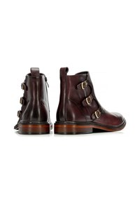 Botas de cuero marrón hasta el tobillo con tres hebillas decorativas, puntera redondeada y tacón de madera apilada. Textura suave y detalles de costura mínimos.