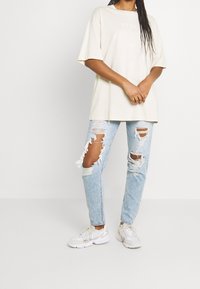 Světle béžové oversized tričko s texturovaným nápisem, v kombinaci s propranými světle modrými džíny s velkými roztrženými místy. Bílé tenisky dokončují vzhled.