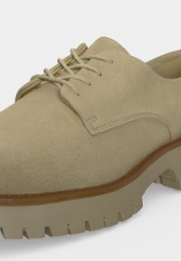 Chaussure beige en suédine à lacets, avec semelle épaisse et crantée et profondes rainures, présentée en gros plan sur fond blanc.