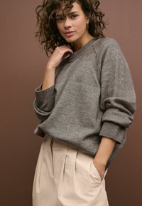 Pulover gri din lână, cu guler și manșete cu creneluri, purtat cu pantaloni bej cu talie înaltă, cu pliuri și buzunare laterale.