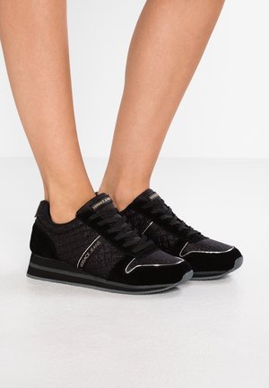 Sorte Versace Jeans-sneakers med sorte snørebånd, båret på bare fødder, med detaljer i ruskind og glitterstof samt en stribet sål.