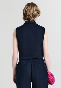 Persona con capelli biondi corti che indossa un top senza maniche blu navy e pantaloni, tenendo un clutch intrecciato rosa brillante dietro la schiena.