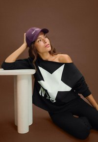 Felpa nera oversize con una grande stella bianca e il testo "NYC", abbinata a un cappellino da baseball viola, su uno sfondo marrone.