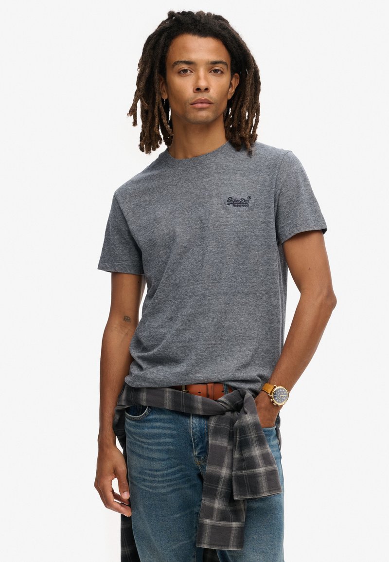 Grijze t-shirt met korte mouwen en ronde hals, gecombineerd met blauwe jeans. Een zwart en grijs geruite blouse om de taille geknoopt, en een detail van een horloge om de pols.