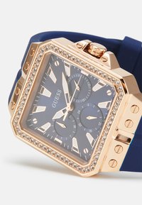 Roségoldene Uhr mit quadratischem Ziffernblatt, blauer Gummiband, funkelnden Kristallen um die Lünette und mehreren Unterzifferblättern auf einem dunkelblauen Hintergrund.