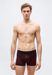 Νέος άνδρας με κατσαρά μαλλιά που φοράει σκούρα βυσσινί μποξεράκια Nike, στέκεται μπροστά σε απλό ανοιχτό γκρι φόντο.