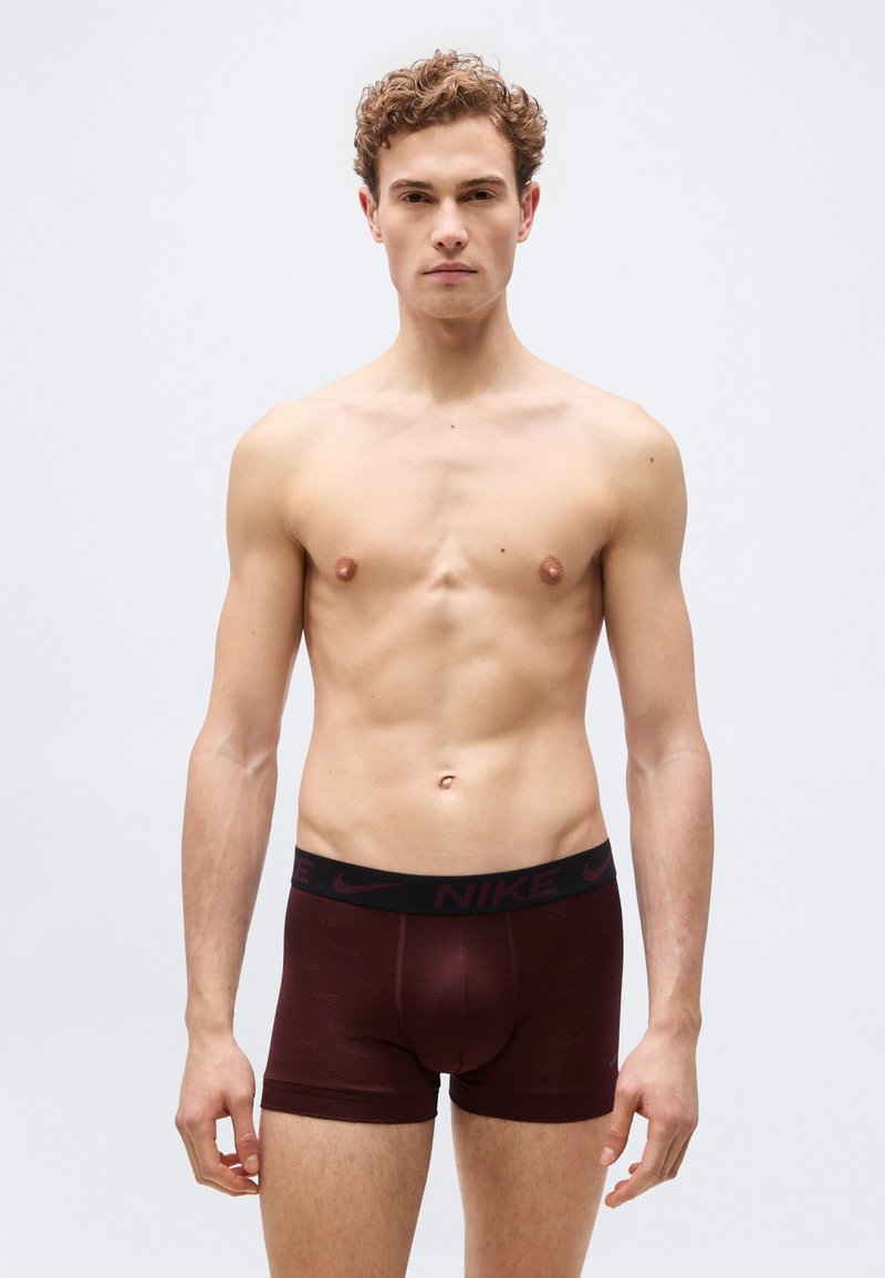 Νέος άνδρας με κατσαρά μαλλιά που φοράει σκούρα βυσσινί μποξεράκια Nike, στέκεται μπροστά σε απλό ανοιχτό γκρι φόντο.