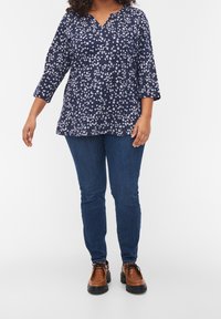 Blusa navy con motivo floreale bianco, scollatura a V, e maniche a tre quarti, abbinata a jeans blu e scarpe stringate marroni.