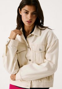 Chaqueta de mezclilla beige con un corte holgado, cierre de botones en la parte frontal, dos bolsillos en el pecho y detalles en botones plateados. Textura suave y diseño casual.