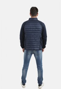 Veste navy avec un devant matelassé, tissu lisse sur les manches, et poignets côtelés. Associée à un jean bleu et des chaussures noires, vue de l'arrière.