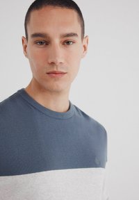 Crewneck-tröja i blått och grått med ett horisontellt randmönster. Tillverkad av mjukt stickat tyg. Inkluderar en liten logotyp på bröstet.