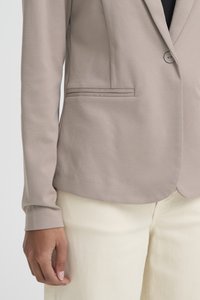 Primer plano de una persona que lleva una blazer beige con un solo botón y pantalones de color crema, mostrando el brazo izquierdo y la mano relajados a un lado.