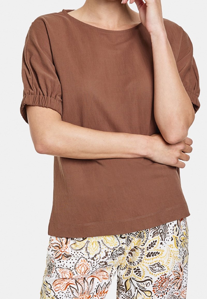 Personne portant un haut marron à manches courtes avec des manches froncées et un pantalon à motif floral, bras croisés et main près du visage.