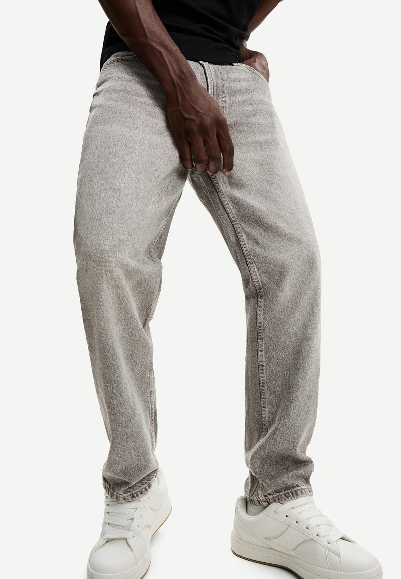 Homme portant un jean ample gris clair, des baskets blanches et une chemise noire, debout avec une main près de la taille et les jambes légèrement écartées.