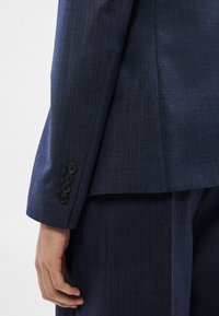 Detailní záběr na osobu v tmavě modrém obleku se čtyřmi černými knoflíky na rukávu, ruka volně podél těla.