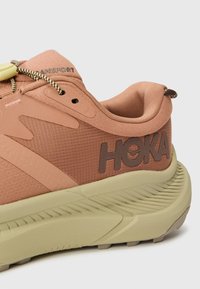 Chaussure de course Hoka dans des tons pêche atténués avec un dessus en mesh texturé, une semelle épaisse beige et un logo distinctif sur le côté. Lacets élastiques inclus.