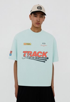 Jonge man die een lichtblauw oversized T-shirt draagt met de tekst "TRACK Fever" en een beige baseballpet tegen een effen achtergrond.